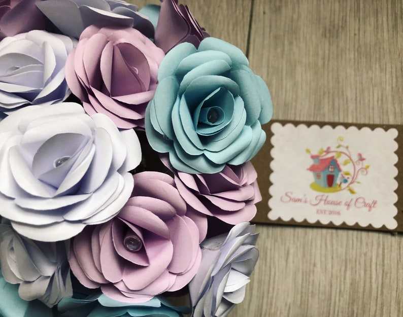 May include: A bouquet of paper flowers in shades of light blue, lavender, and white. The flowers are arranged in a circular shape and have a delicate, layered appearance. The flowers are accented with small, clear beads in the center of each bloom.