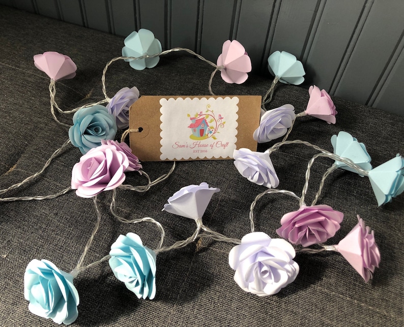 May include: A string of fairy lights with paper flower accents in shades of pink, blue, and white. The lights are strung together with a clear wire and have a small brown tag with the text "Sara's House of Craft Est 2018".