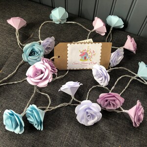May include: A string of fairy lights with paper flower accents in shades of blue, purple, and pink. The lights are strung together with a clear wire. The flowers are made of paper and have a delicate, ruffled appearance.