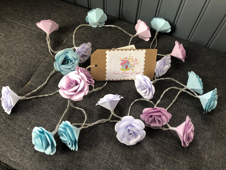 May include: A string of fairy lights with paper flower accents in shades of blue, purple, and pink. The lights are strung together with a clear wire. The flowers are made of paper and have a delicate, ruffled appearance.