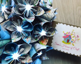 May include: A bouquet of origami flowers made from maps with white lights. The flowers are blue and white and the maps are printed with black text. The bouquet is sitting on a wooden surface. The text on the map reads "Sam's House of Craft EST 2016".
