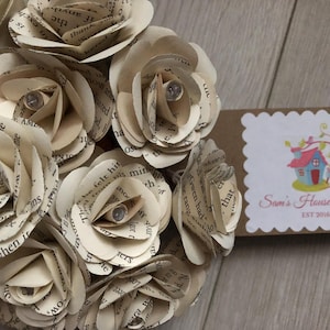 以下が含まれることがあります： 古い本のページで作られた紙のバラのブーケ。バラはクリーム色で、ヴィンテージな雰囲気があります。ブーケは、ブラウンの表面に置かれ、小さな看板には「Sam's House of Craft EST 2016」と書かれています。