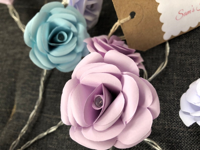May include: A string of fairy lights with paper flower accents. The flowers are in shades of light blue, lavender, and white. The lights are strung on a silver wire.