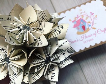 May include: A bouquet of origami flowers made from sheet music. The flowers are white with black musical notes printed on them. The flowers are arranged in a cluster and have a small, clear rhinestone in the center of each flower. The bouquet is sitting on a wooden surface. The text "Sam's House of Craft EST 2016" is visible in the background.