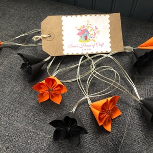 May include: A string of lights with orange and black origami paper flowers. The lights are strung on a clear wire. The flowers are in the shape of a bell. The tag says "Sam's House of Craft Est 2018".
