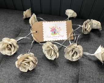 May include: A string of fairy lights with paper roses made from sheet music. The lights are strung on a clear wire and the roses are a cream color. The tag says "Sam's House of Craft Est 2018".
