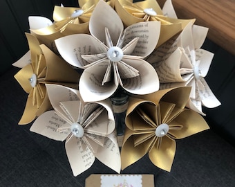 May include: A bouquet of paper flowers made from book pages. The flowers are in shades of white and gold and are arranged in a vase. The flowers are decorated with small white buttons. The bouquet is sitting on a dark surface. The tag on the bouquet reads "Sam's House of Craft".
