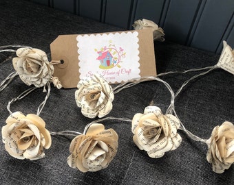May include: A string of fairy lights with paper roses made from old book pages. The lights are strung together with a brown wire. The roses are a cream color and have a vintage look. The tag says "So Homely House of Craft Est 2016".