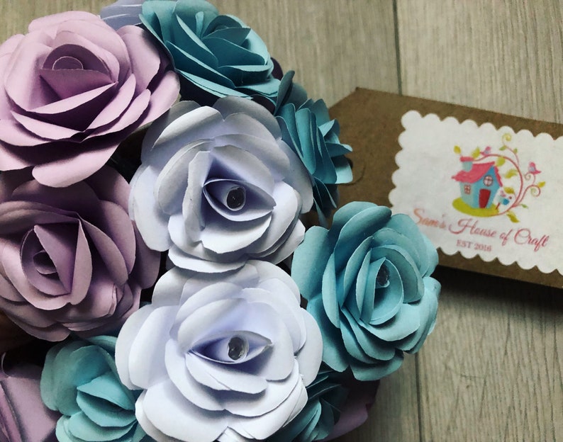 May include: A bouquet of paper flowers in shades of purple, blue, and white. The flowers are arranged in a circular shape and have a delicate, layered appearance. The bouquet is accented with a small, brown tag that reads "Sam's House of Craft Est 2018."