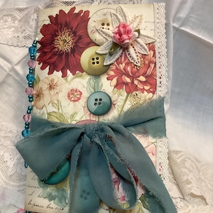 May include: A handmade journal with a floral design featuring red and white flowers. Adorned with decorative buttons, a teal ribbon, and a beaded strand. The journal's edges are trimmed with lace.