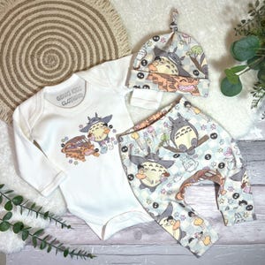 Peut inclure: Un ensemble de vêtements pour bébé composé d'un body blanc à manches longues, d'un pantalon à motifs et d'un bonnet noué. L'ensemble est orné de personnages de dessins animés bruns, gris et bleus. Le body porte l'inscription "GENKI KIDS CLOTHING".