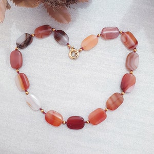 May include: A red, orange, and white carnelian stone necklace with a gold clasp. The necklace is made of oval-shaped stones and is strung on a red cord.