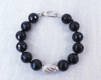 Può includere: Bracciale con perline di onice nero sfaccettate, con chiusura argentata e un elemento decorativo in argento e cristalli. Le perline rotonde e grandi creano un gioiello elegante.