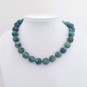 May include: A turquoise beaded necklace displayed on a white mannequin. The necklace features large, round beads with a mottled pattern of teal and dark green. The beads are strung on a light blue cord, creating a striking contrast.