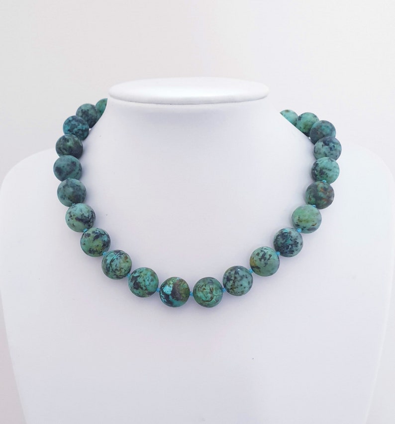 May include: A turquoise beaded necklace displayed on a white mannequin. The necklace features large, round beads with a mottled pattern of teal and dark green. The beads are strung on a light blue cord, creating a striking contrast.