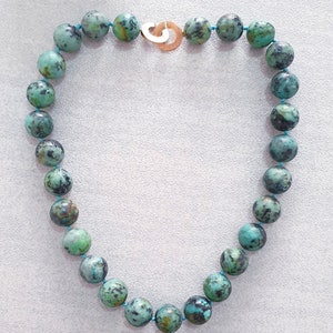 May include: A turquoise beaded necklace with a gold-colored clasp. The beads are round, with a mottled pattern of green and black. The necklace is strung on a thin, light blue cord. The clasp is a simple, open design.