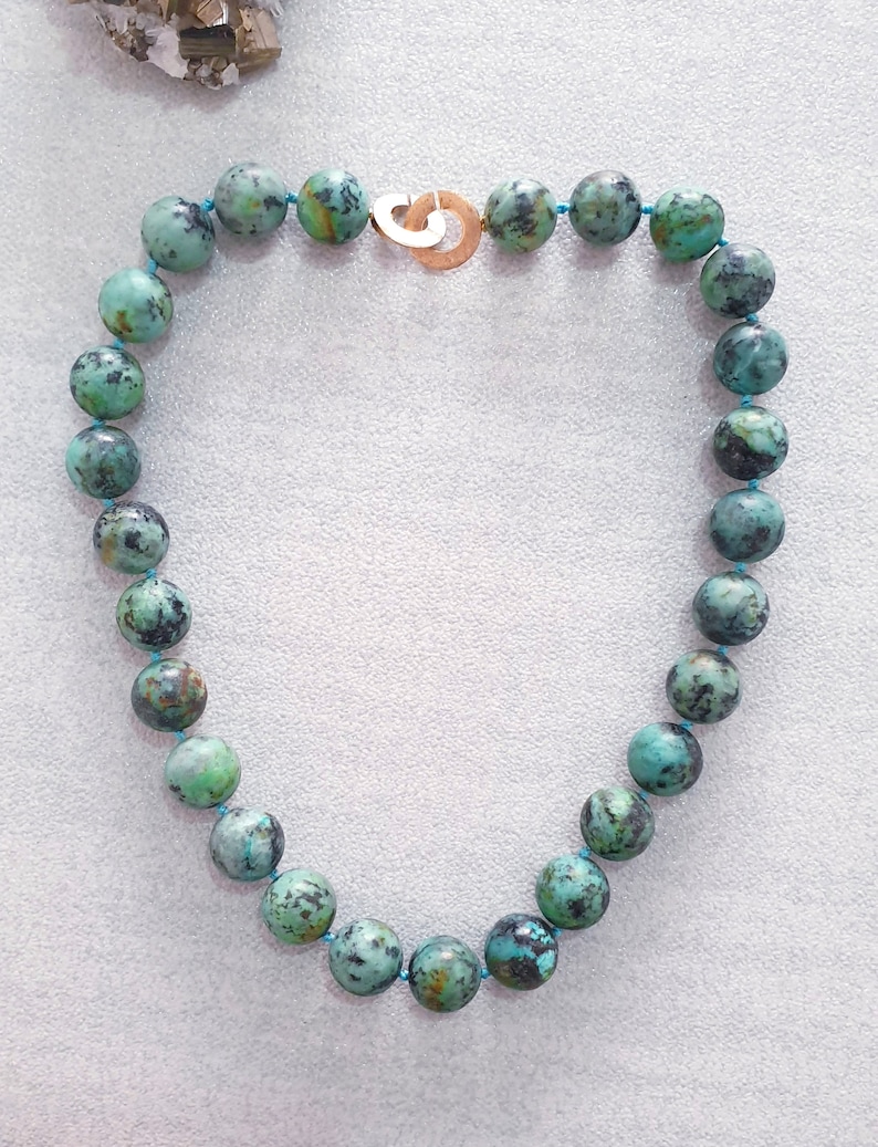 May include: A turquoise beaded necklace with a gold-colored clasp. The beads are round, with a mottled pattern of green and black. The necklace is strung on a thin, light blue cord. The clasp is a simple, open design.