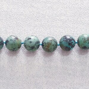 May include: Close-up of a strand of turquoise gemstone beads. The beads are round, with a mottled pattern of green and dark blue. They are strung on a thin, light blue cord, creating a necklace or bracelet.
