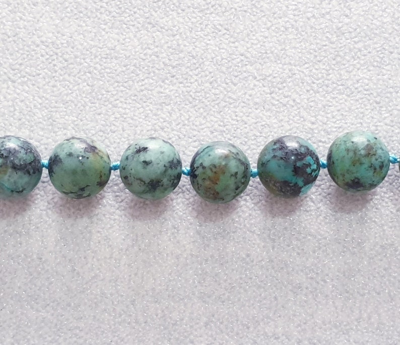May include: Close-up of a strand of turquoise gemstone beads. The beads are round, with a mottled pattern of green and dark blue. They are strung on a thin, light blue cord, creating a necklace or bracelet.