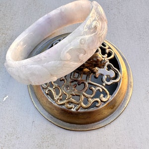 May include: A white jade bangle with intricate carved floral designs. The bangle is resting on a decorative metal stand.