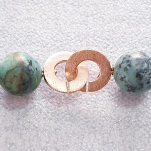 May include: Close-up of a necklace featuring large, round turquoise beads with dark green and brown marbling. The necklace has a gold-colored clasp with interlocking circles and is strung on a light blue cord.
