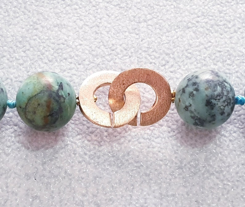 May include: Close-up of a necklace featuring large, round turquoise beads with dark green and brown marbling. The necklace has a gold-colored clasp with interlocking circles and is strung on a light blue cord.