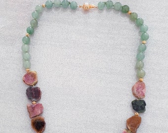 Könnte beinhalten: Eine Halskette mit grünen Perlen und grob geschnittenen Turmalinsteinen in Rosa-, Braun- und Grüntönen. Die Halskette ist mit goldfarbenen Akzenten und einem goldenen Verschluss versehen. Der Schmuck wird auf hellem Hintergrund mit rosa Blüten präsentiert.