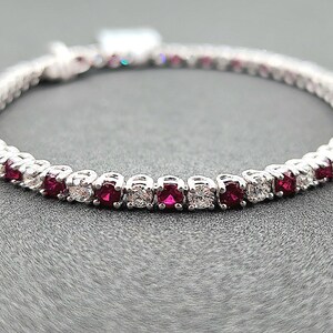 May include: A tennis bracelet featuring alternating red and clear gemstones set in a silver-colored metal. The bracelet is displayed on a gray surface, showcasing its elegant design and sparkle. This piece of jewelry is a classic accessory.