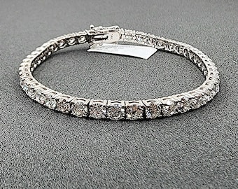 May include: A silver tennis bracelet with numerous round-cut, clear gemstones. The bracelet is clasped and rests on a dark gray surface. A small white tag is attached to the clasp.