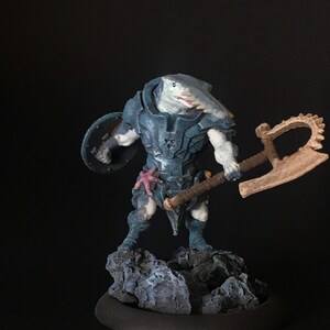May include: A blue and white painted miniature figurine of a shark-like creature wearing armor and holding a large axe and shield. The figurine is standing on a rocky base.