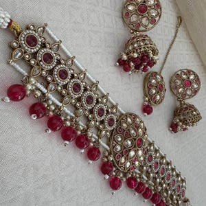 May include: A gold-tone choker necklace with red and white stones, a matching pair of earrings, and a matching nose ring. The necklace has a layered design with multiple strands of pearls and red stones.