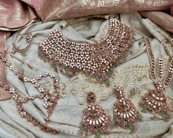 May include: An assortment of rose gold-toned jewelry, including a statement necklace, earrings, a bracelet, and a nose ring, all adorned with reflective mirrored accents and delicate green and pink beads. The pieces are arranged on a cream-colored fabric.