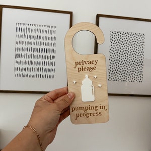 Pu&ograve; includere: Appendiporta in legno con il testo "privacy please" e "pumping in progress". Al centro del cartello &egrave; presente una sagoma bianca di un biberon.