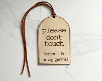 Può includere: Un'etichetta di legno con un cordino in pelle marrone. L'etichetta ha la forma di un arco e ha il testo "please don't touch i'm too little for big germs!"