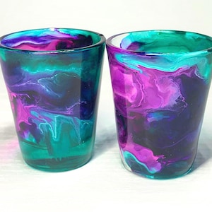 May include: Two shot glasses with a colorful, abstract design. The glasses feature a blend of teal, purple, and dark blue hues, creating a marbled effect. The shot glasses are made of clear glass.