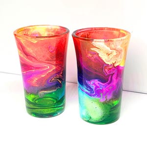 May include: Two shot glasses with vibrant, swirling colors. The glasses feature a blend of red, pink, purple, green, and yellow hues, creating a marbled effect. The base of each glass is green, transitioning to a mix of colors towards the top.