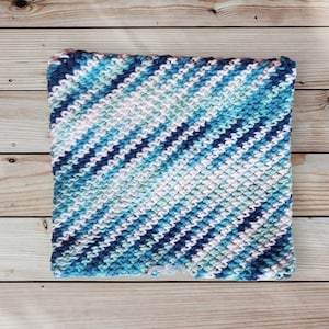 May include: A square, hand-knitted washcloth with a diagonal striped pattern. The washcloth features a blend of blue, white, and light green yarns. The texture of the knitted fabric is clearly visible, with each stitch creating a raised pattern.