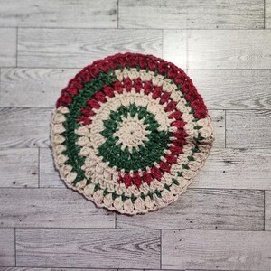 May include: A handmade, circular crochet item in a festive color scheme. The design features concentric rings of red, green, and cream-colored yarn. The item is likely a coaster or trivet, adding a touch of handmade charm to any space.