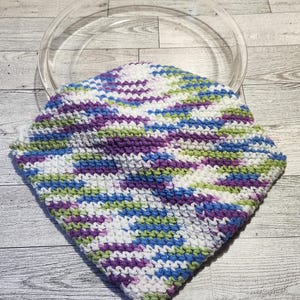 Może przedstawiać: Kolorowy, ręcznie szydełkowany podkładka pod garnek z przezroczystym, okrągłym uchwytem. Podkładka ma geometryczny wzór w odcieniach fioletu, niebieskiego, zielonego i białego. Kwadratowy kształt jest idealny do ochrony powierzchni przed gorącymi przedmiotami.