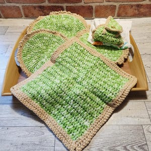 Peut inclure: Un ensemble de linge de cuisine crocheté à la main comprenant une manique carrée, deux dessous de verre ronds et une petite pochette pliée, réalisés en fil vert clair et blanc nuancé avec des bordures beige, disposés sur un plateau en bois.