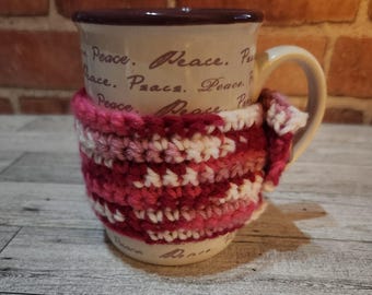 May include: A beige ceramic mug with the word "Peace" printed repeatedly, wrapped in a handmade crochet cozy. The cozy features a striped pattern in shades of pink, white, and burgundy, adding a touch of warmth.