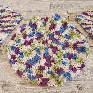 Puede incluir: Tres artículos de ganchillo hechos a mano. Se muestran dos agarraderas cuadradas y un salvamanteles redondo. El patrón de ganchillo utiliza hilo blanco, morado, verde y azul. Los artículos están dispuestos sobre una superficie de madera clara.