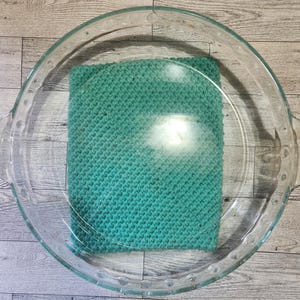May include: A teal knitted washcloth, submerged in a clear glass pie dish filled with water. The washcloth is square and has a textured surface. The pie dish has a scalloped edge and is on a light-coloured wooden surface.