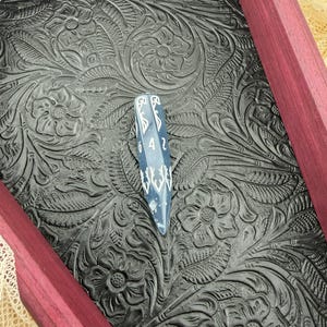 May include: A long, slender, dark blue polyhedral die with white numbering and decorative white filigree patterns, resting on a black tray featuring an embossed floral pattern and framed by deep purple wooden edges.