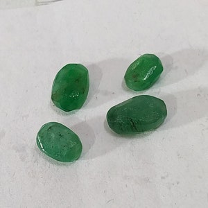 May include: Four green emerald gemstones, each with a slightly different shape and size, are displayed on a white surface.