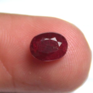 Puede incluir: Gema de rubí facetada de forma ovalada. La gema de color rojo intenso se sostiene entre los dedos, mostrando su superficie pulida y su estructura interna. Las facetas del rubí capturan la luz, creando un efecto brillante. Vista de cerca.