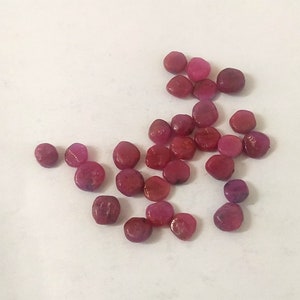 Pode incluir: Uma coleção de pequenas pedras preciosas redondas, cor-de-rosa e vermelhas. As pedras preciosas estão espalhadas sobre uma superfície branca.