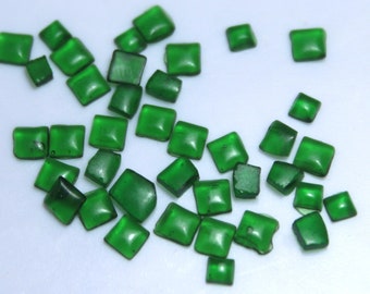 May include: A collection of small, square, green gemstones. The gemstones are scattered on a white surface.