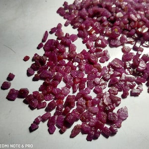 Op de afbeelding: Een hoop kleine, ruwe, roze robijn edelstenen. De robijnen zijn van verschillende vormen en maten en zijn verspreid over een wit oppervlak.