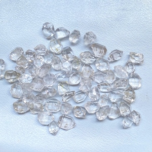 Op de afbeelding: Een verzameling van heldere kwartskristallen, ook wel Herkimer-diamanten genoemd, in verschillende vormen en maten. De kristallen zijn in een cluster op een lichtblauw oppervlak gerangschikt.
